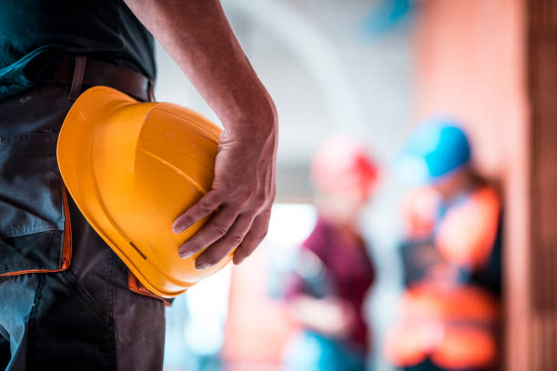 Architects are using digital tablet on construction site. Manual worker is in front and holds helmet in his hand.