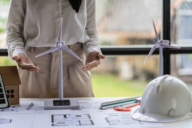 Young Asian architect with blueprints working on lapotp in office. Career working hard on wooden table, painting with windmills, designing model of renewable wind house building. and sunlight