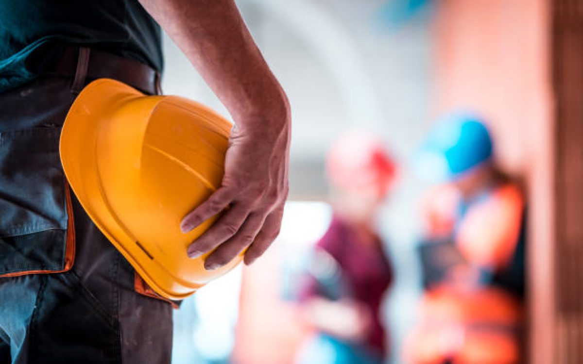 Architects are using digital tablet on construction site. Manual worker is in front and holds helmet in his hand.