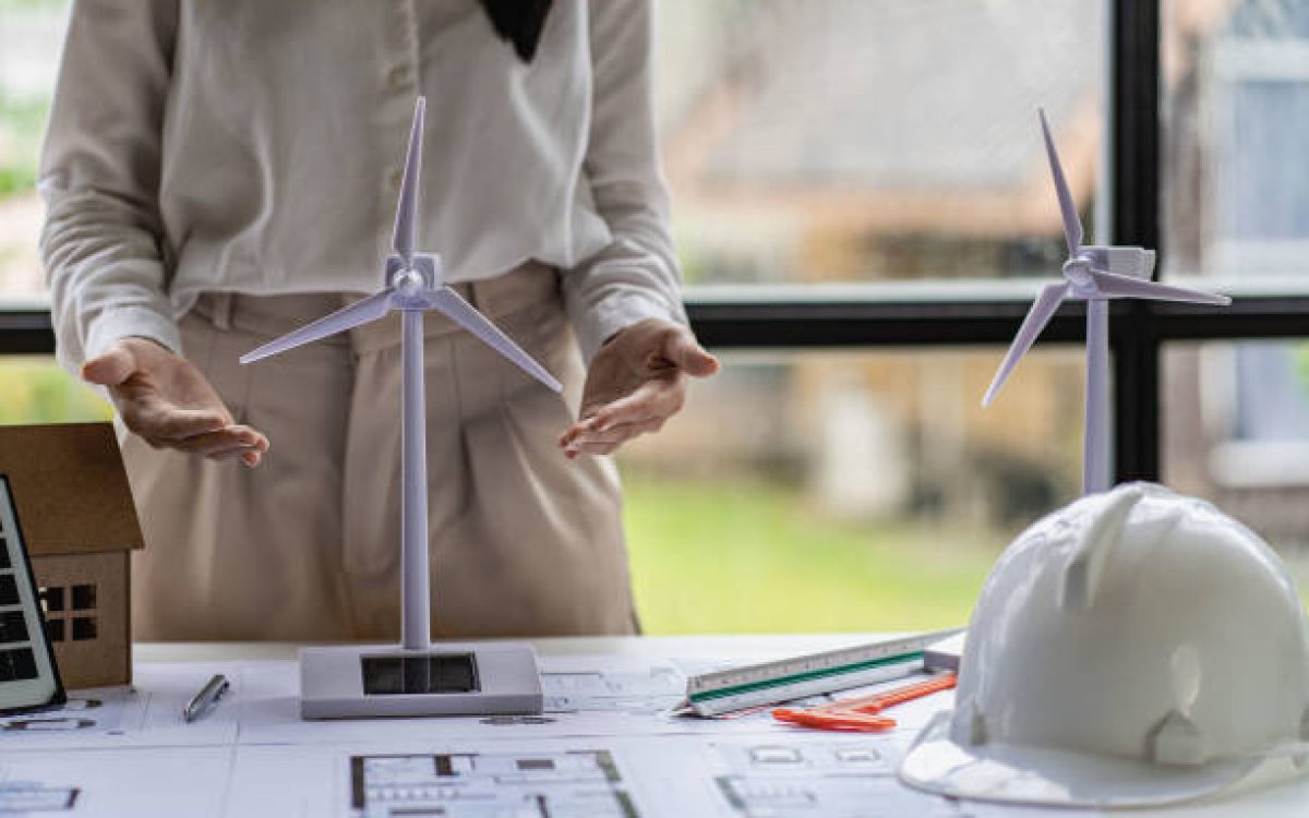 Young Asian architect with blueprints working on lapotp in office. Career working hard on wooden table, painting with windmills, designing model of renewable wind house building. and sunlight