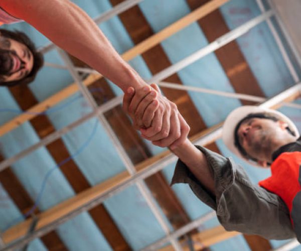 Two construction workers in safety gear engage in a firm handshake, symbolizing teamwork and agreement on a construction site.