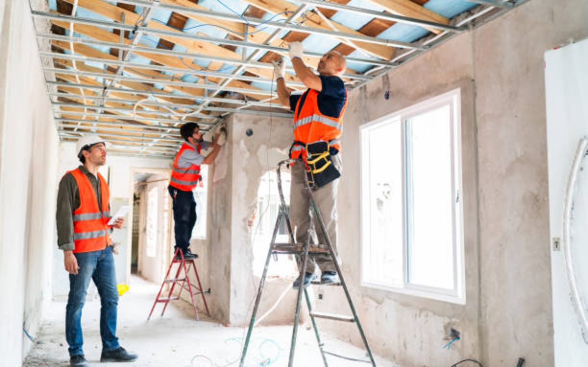 Three construction workers in safety vests and helmets work together installing ceiling framework and windows during an interior renovation project.